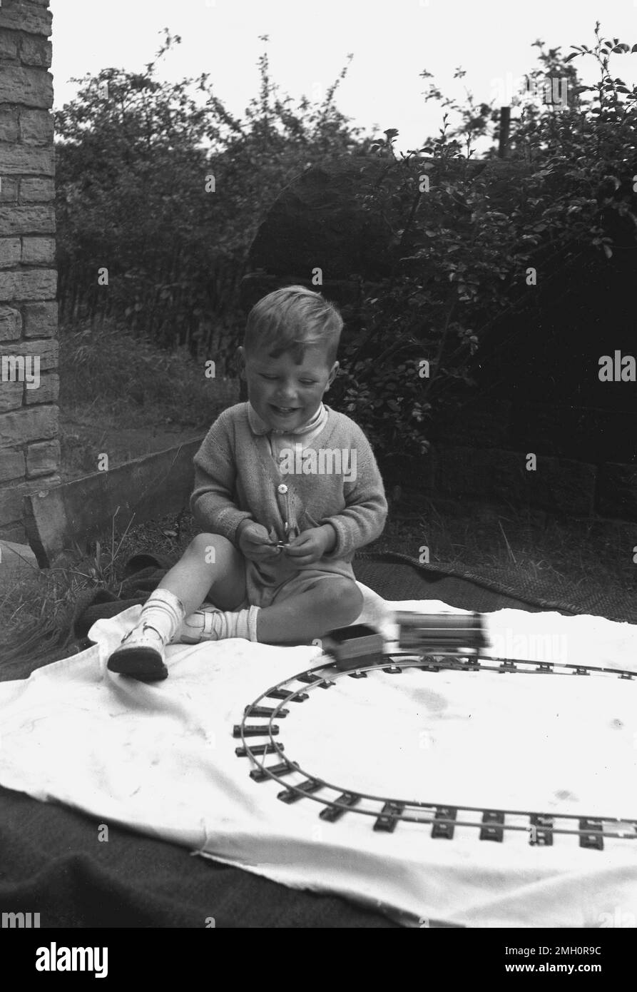 1960s, historical, young boy sitting on a sheet on a rug outside, playing with his train set, England, UK. A toy locomotive on the railtrack pulling a wagon. Stock Photo