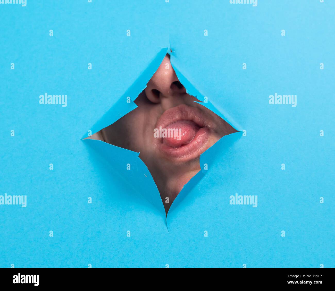 Woman sticking her tongue out of a hole on blue paper background.  Stock Photo