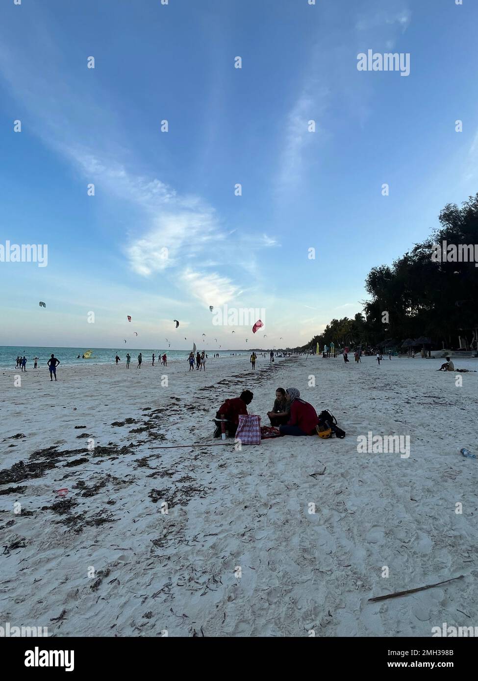 Many people use kites in front of the beach, and one family sits on the white sand watching the beach in Zanzibar, Tanzania. Stock Photo