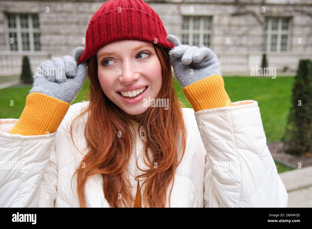 Close up portrait of beautiful redhead woman in red knitted hat, warm gloves, smiling and looking happy at camera, sitting in park Stock Photo