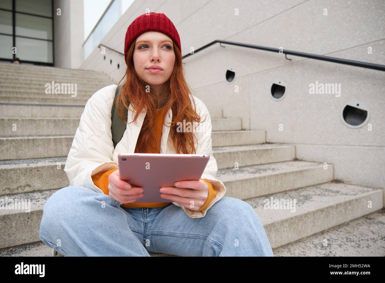 Young stylish girl, redhead female students sits on stairs outdoors with digital tablet, reads, uses social media app on gadget, plays games while Stock Photo