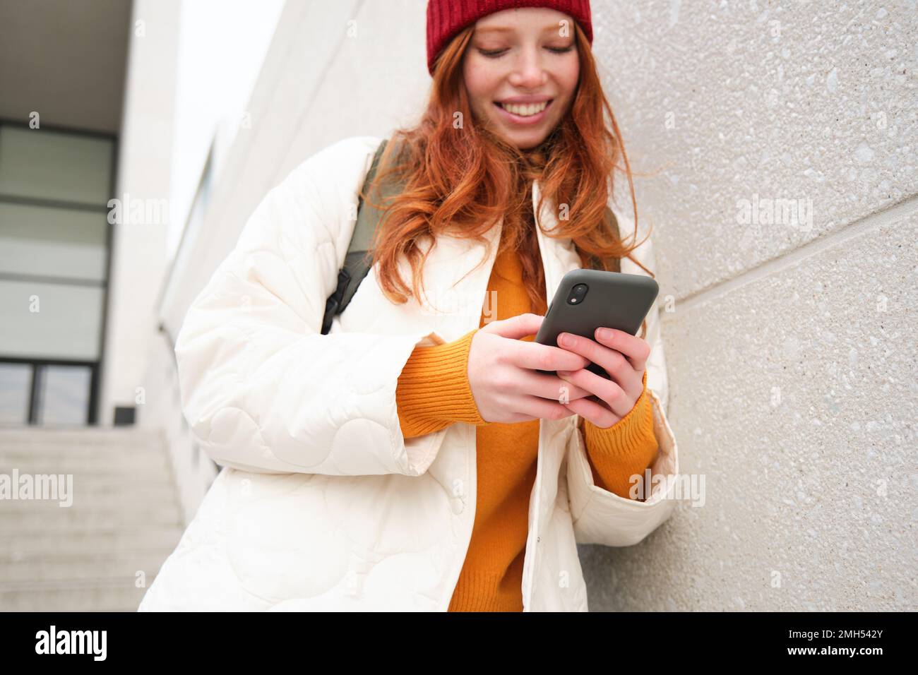 Redhead girl, young woman tourist with backpack, holds smartphone, looks for route on mobile application, searches for hotel on phone map, smiles Stock Photo