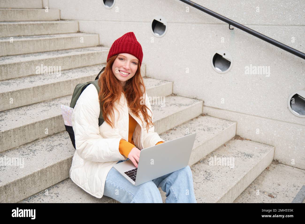 Portrait of young girl traveller, sitting with backpack and map of city, working on laptop, connecting to public wifi and sitting on stairs outdoors Stock Photo