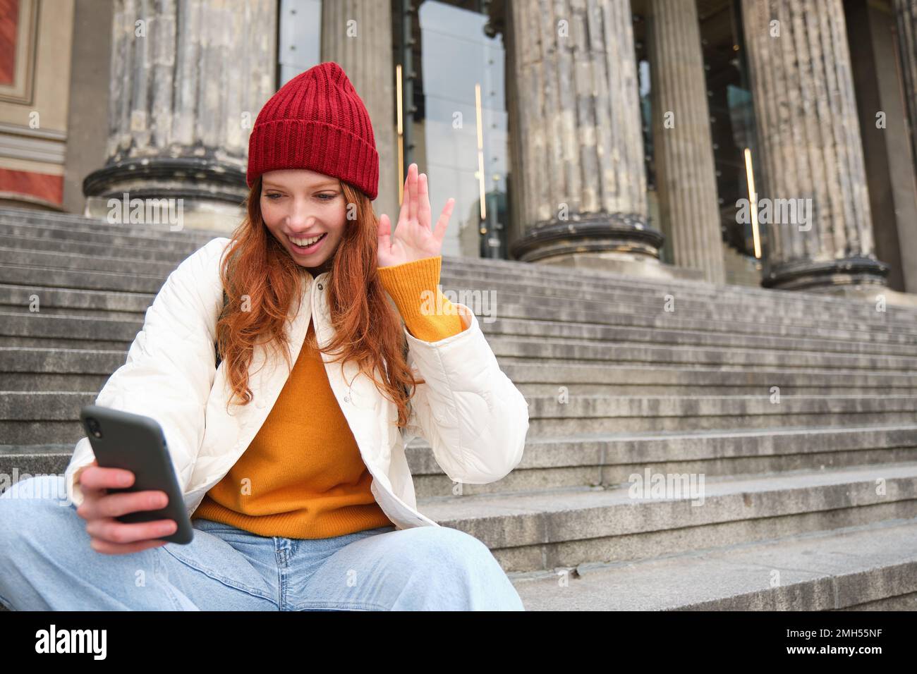 Young redhead woman sits on stairs outdoors and waves hand at smartphone camera, video chats with friends, connects to public wifi Stock Photo