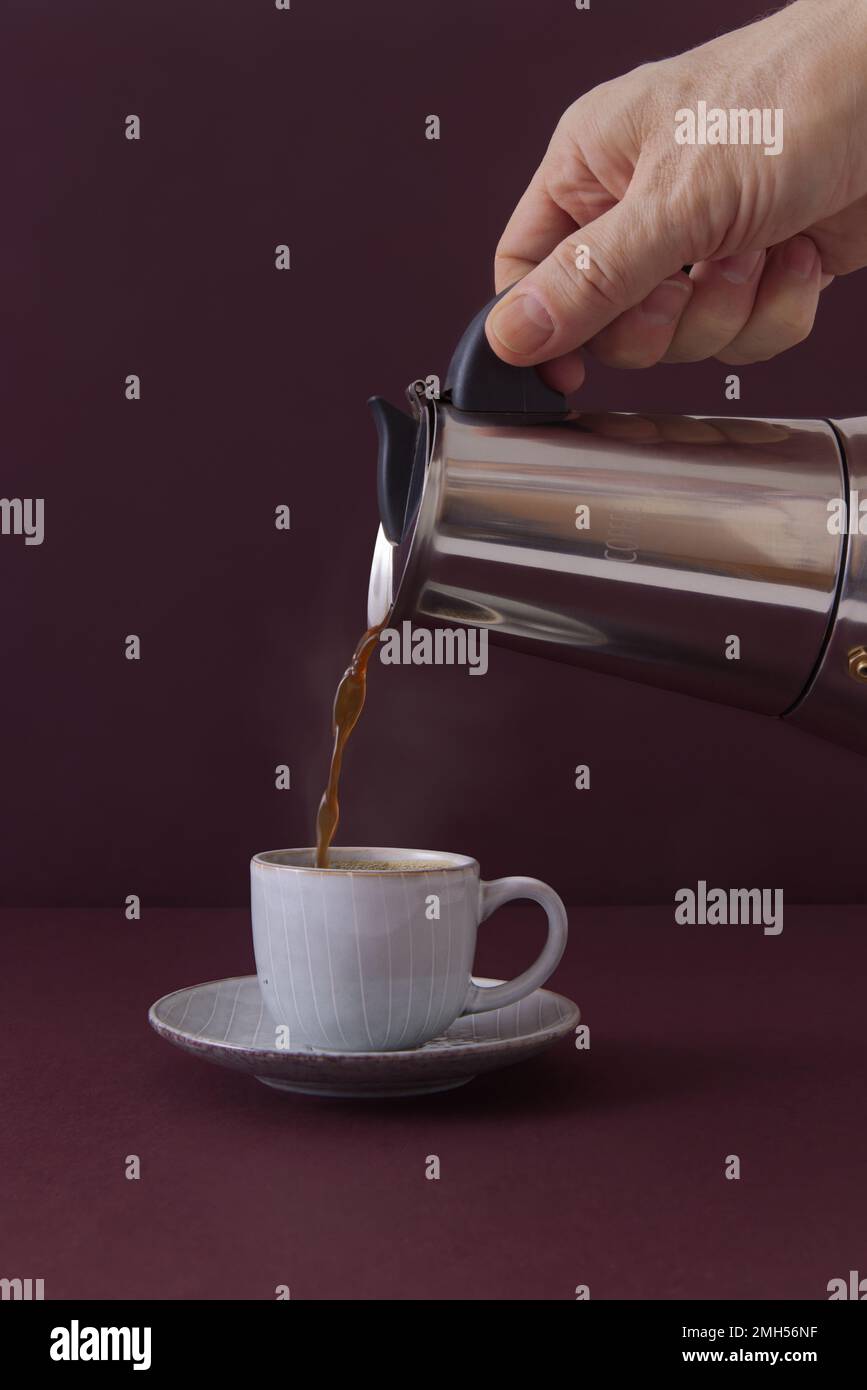 A hand pouring coffee from a mocha pot into a small cup next to a pile of coffee beans against a dark burgundy background. Portrait orientation, no AI Stock Photo