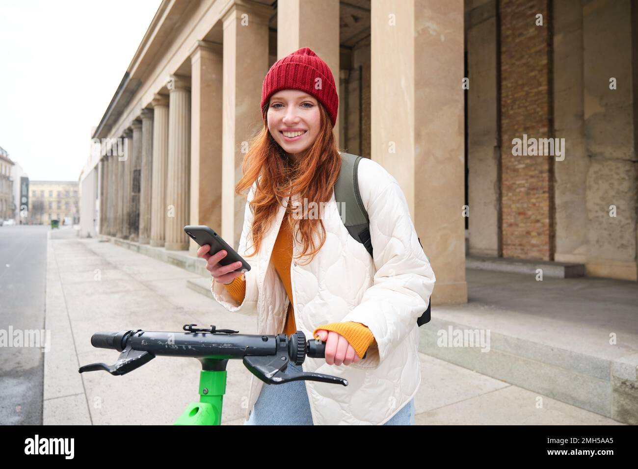 Beautiful young woman, tourist with backpack, rents e-scooter to travel around town, uses mobile phone application Stock Photo