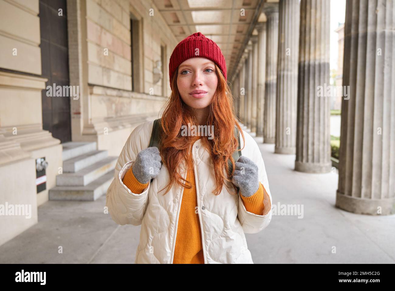 Smiling redhead girl with backpack, walks in city and does sightseeing, explore popular landmarks on her tourist journey around Europe Stock Photo