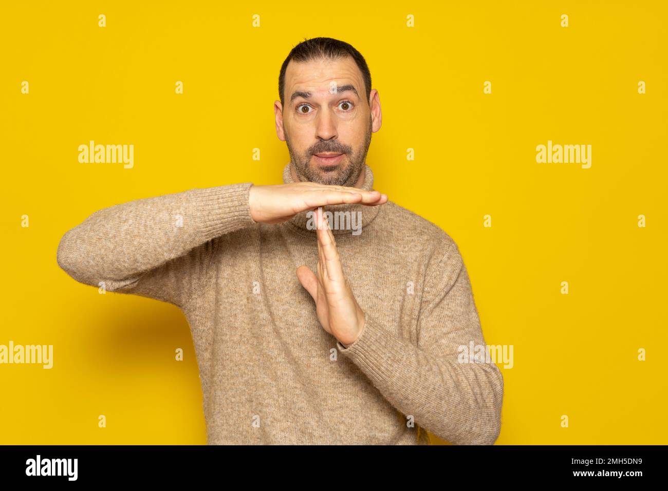 Bearded hispanic man wearing a turtleneck sweater showing time out gesture, tired of a lot of work. Indoor studio shot isolated on yellow background. Stock Photo