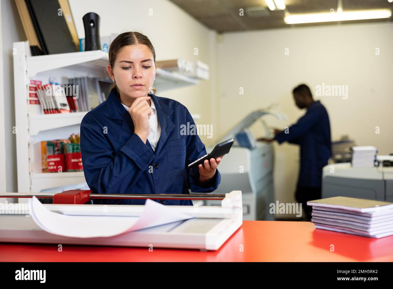 Focused young female typographer in uniform calculates the cost of printed notepads and sheet in the typography Stock Photo