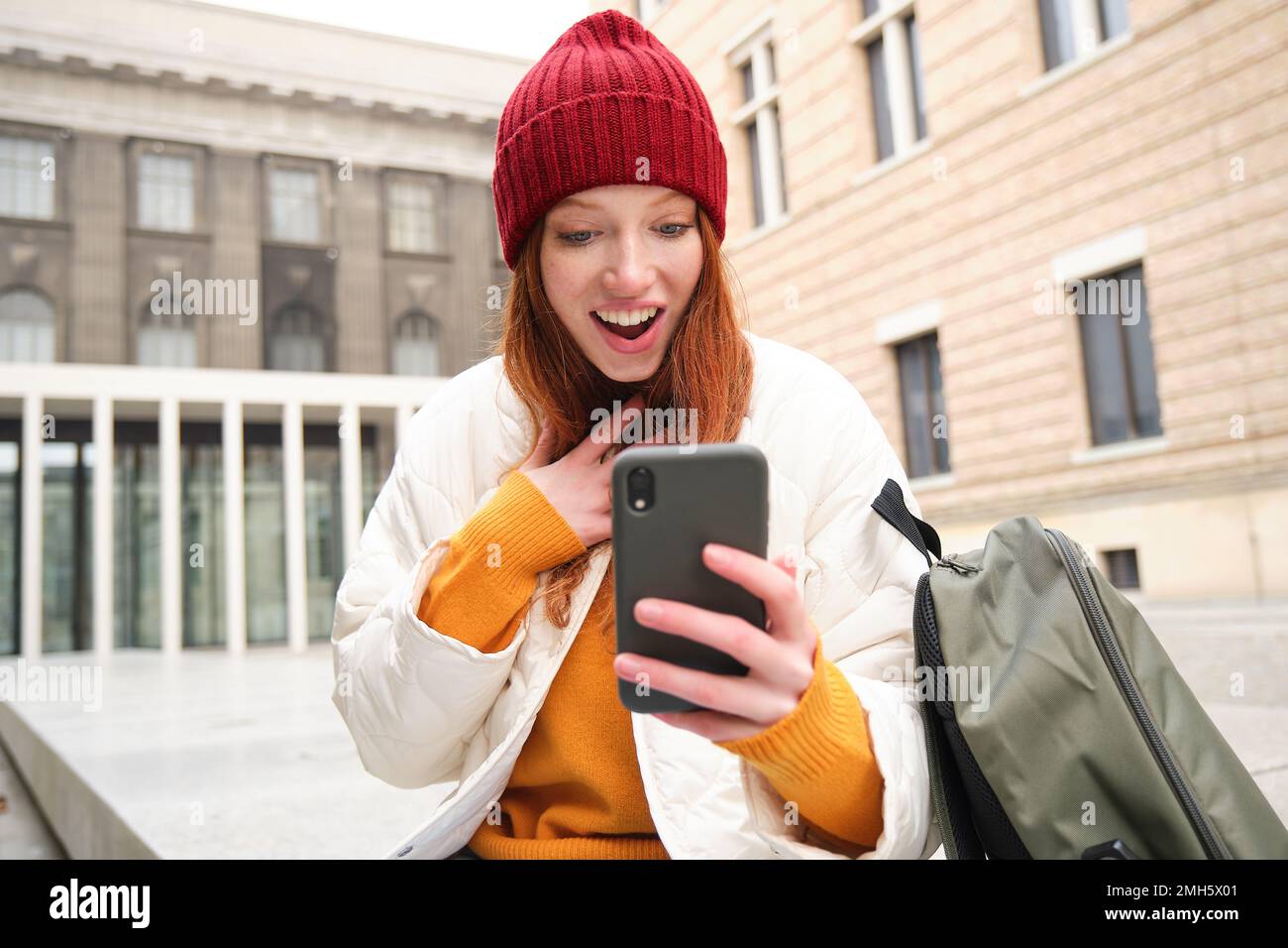 Young redhead woman with smartphone, sitting outdoors with backpack, student looking at her mobile phone, texts message Stock Photo