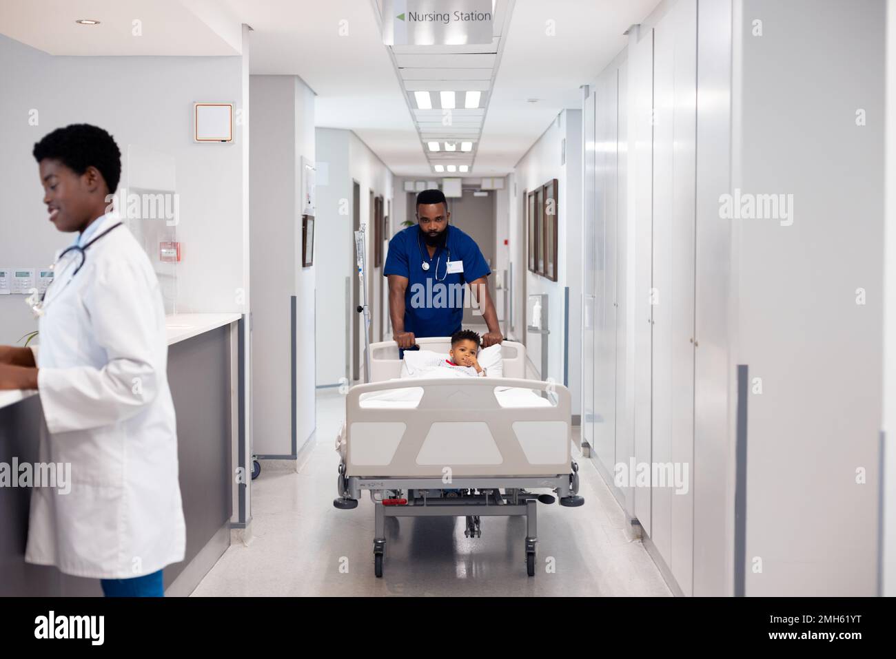 African american male doctor pushing boy patient in hospital bed in busy corridor with copy space Stock Photo