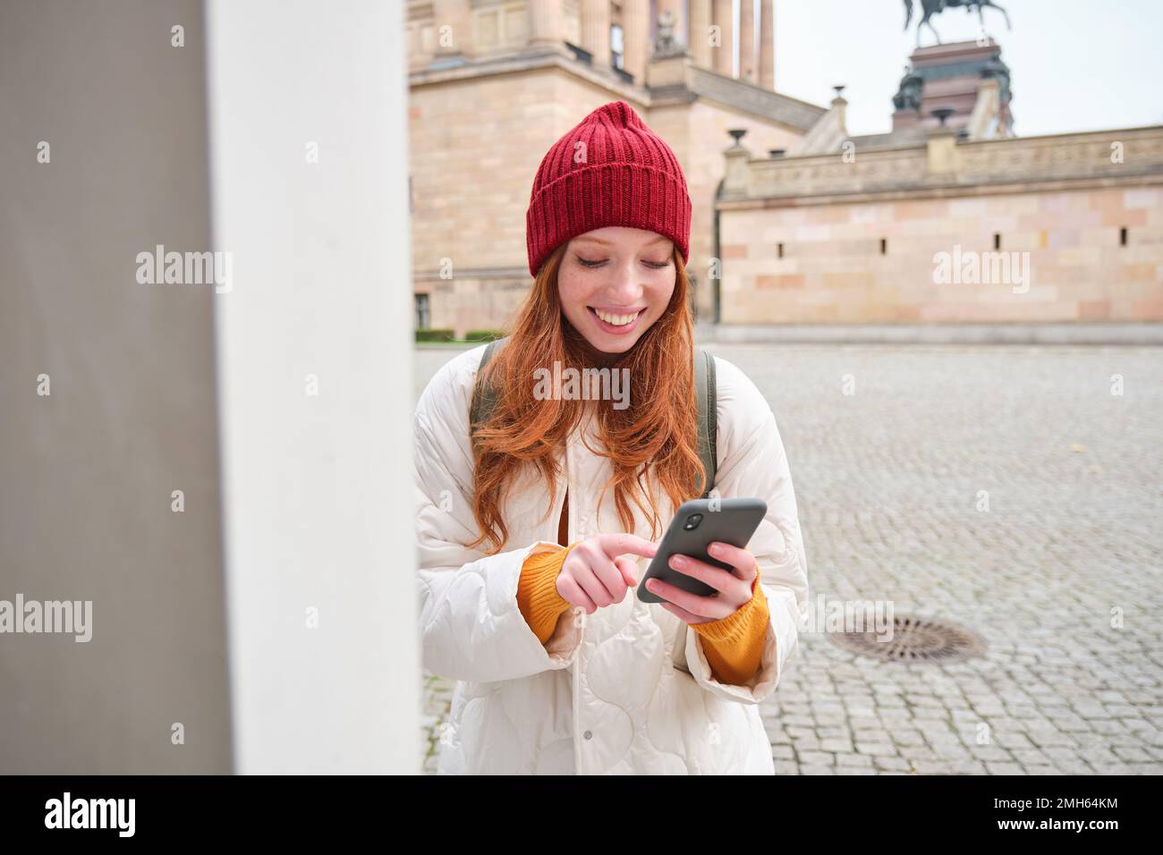 Smiling redhead girl tourist, walks around city and explores popular landmarks, sightseeing, holding smartphone, looking at her mobile app and checks Stock Photo