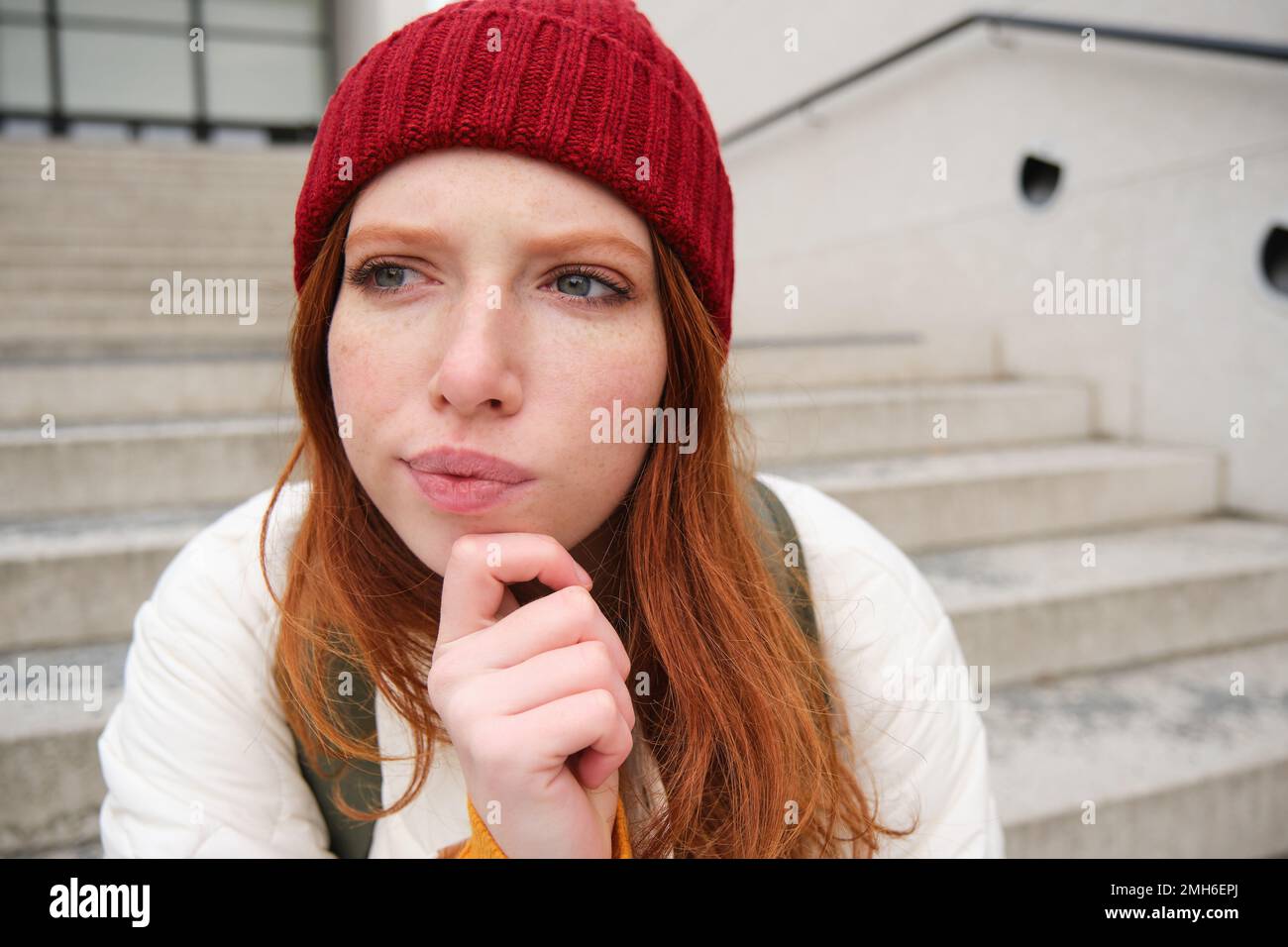Portrait of redhead girl in red hat, looks thoughtful, touches her chin, frowns and thinks with complicated face expression, sits on stairs outdoors Stock Photo