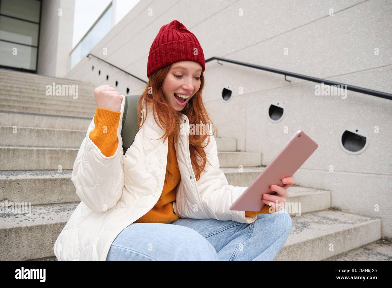 Happy stylish redhead girl, student in red hat, holds digital tablet, uses social media app, searches something online, connects to wifi Stock Photo