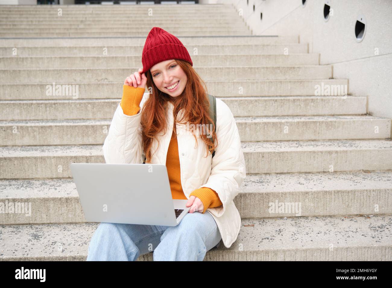 Smiling redhead girl, young woman typing on laptop keyboard, sitting outdoors on stairs with computer, working remote, doing her homework on fresh air Stock Photo