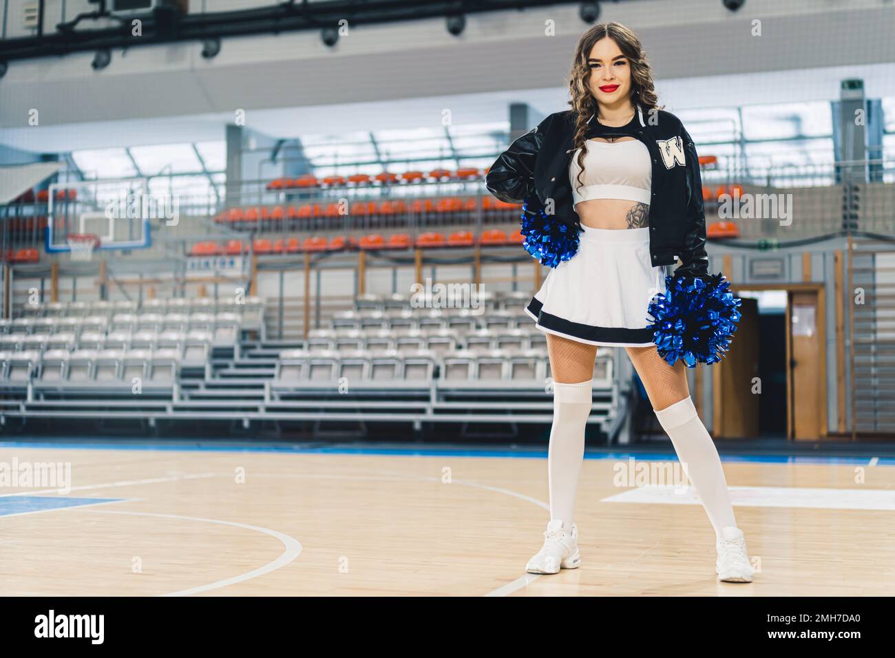 Full shot of a cheerleader in uniform with a jacket posing on basketball court. Sports hall's boxes blurred in the background. High quality photo Stock Photo