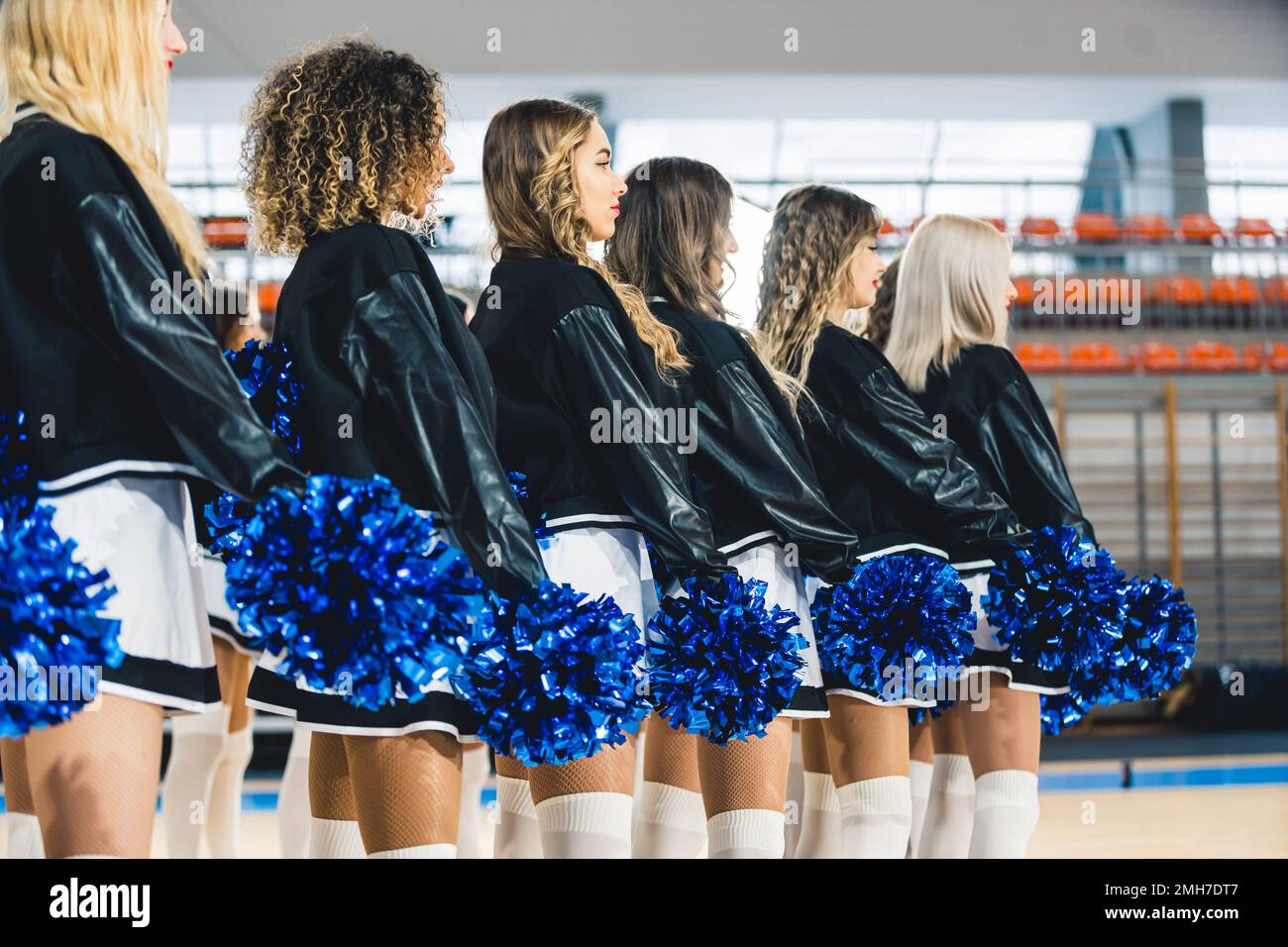 Side view of cheerleaders in uniforms standing in line with blue pom-poms in their hands. Blurred sports hall in the background. High quality photo Stock Photo
