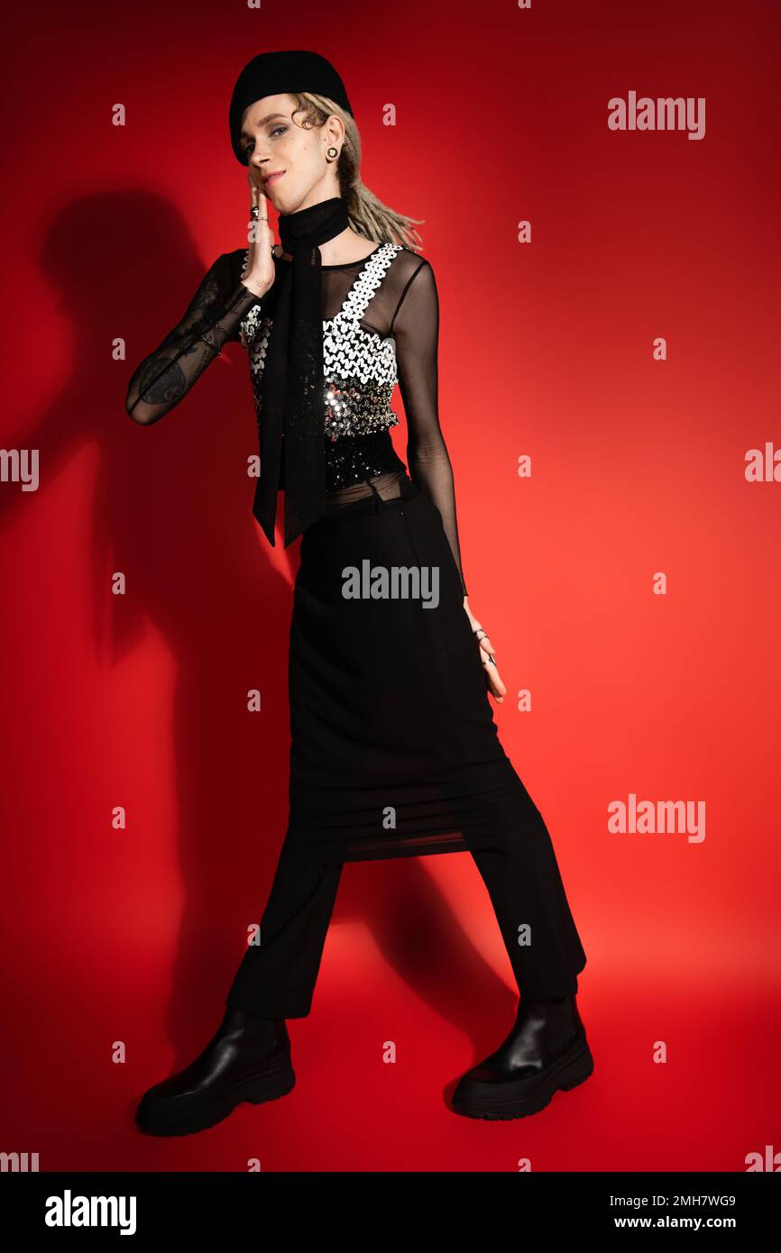 full length of smiling nonbinary person in black elegant clothes holding hand near face on red background with shadow,stock image Stock Photo