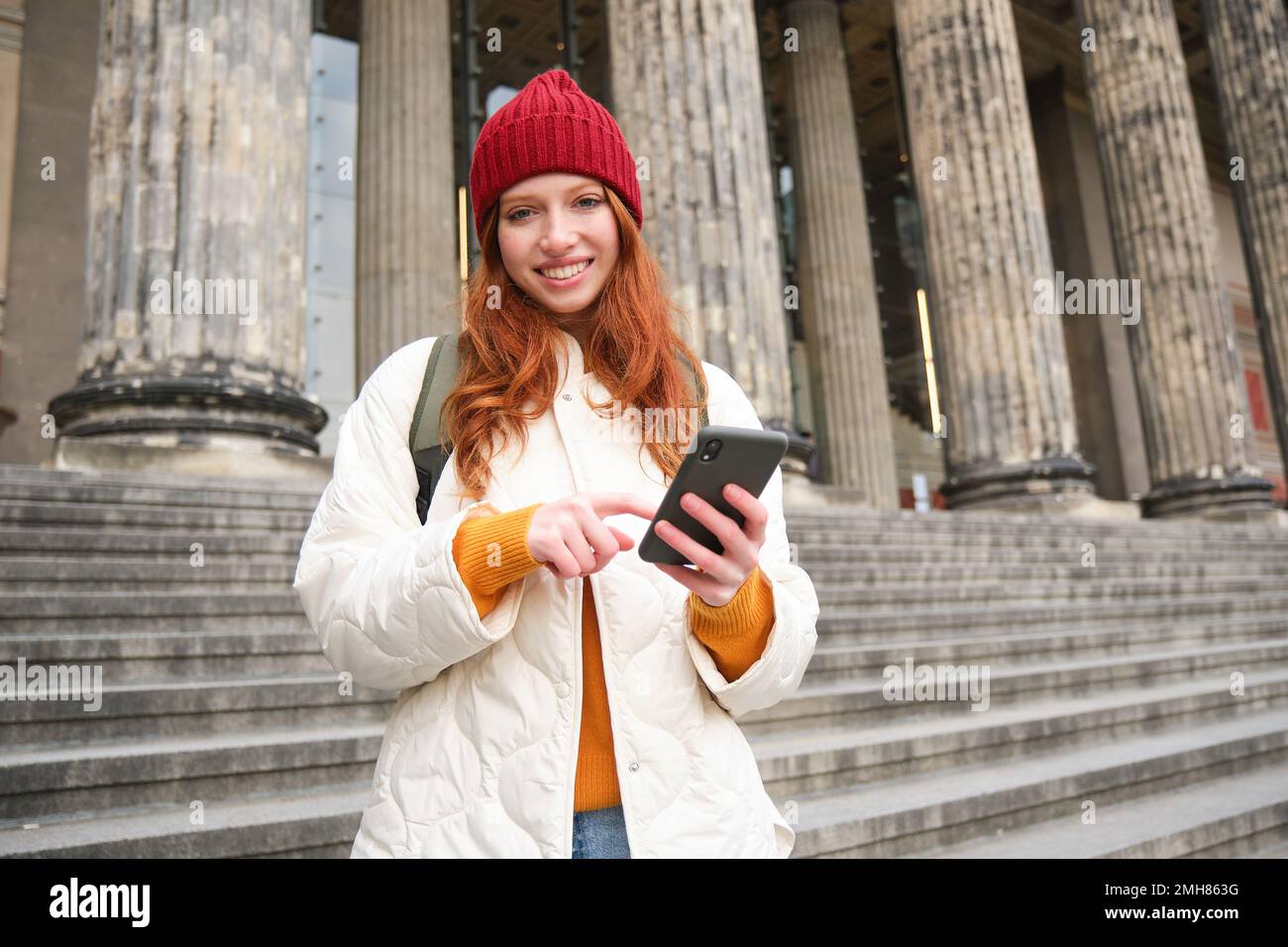 Portrait of young redhead woman holding mobile phone, using smartphone app, walking in city centre, using map application or texting a message Stock Photo