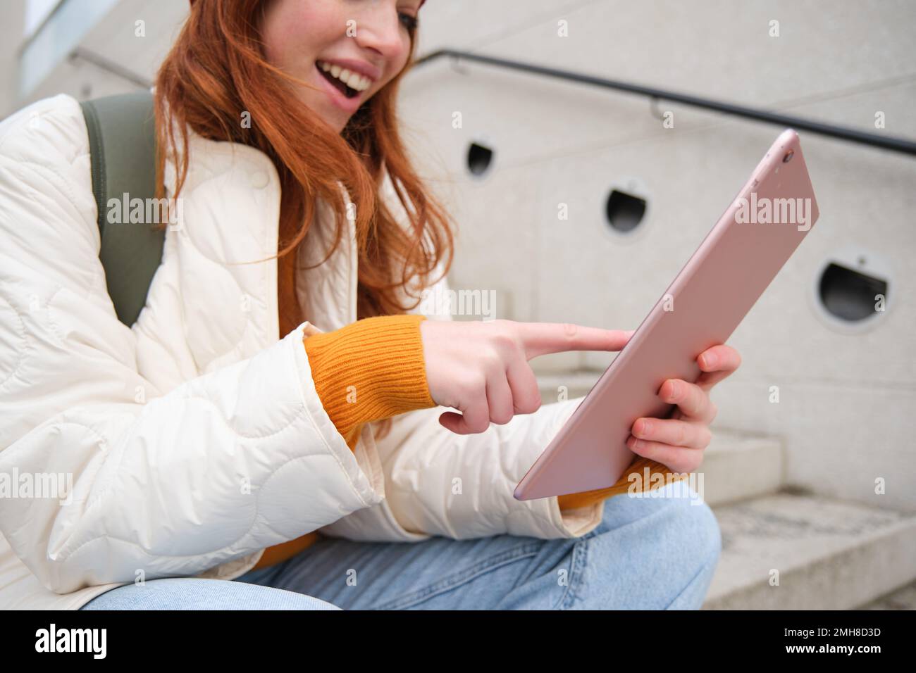 Young stylish girl, redhead female students sits on stairs outdoors with digital tablet, reads, uses social media app on gadget, plays games while Stock Photo
