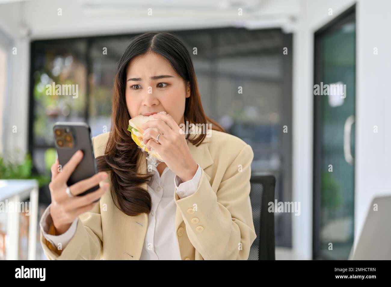 Hungry and busy millennial Asian businesswoman eating a sandwich while replying or checking her clients email on the phone. busy lifestyle concept Stock Photo