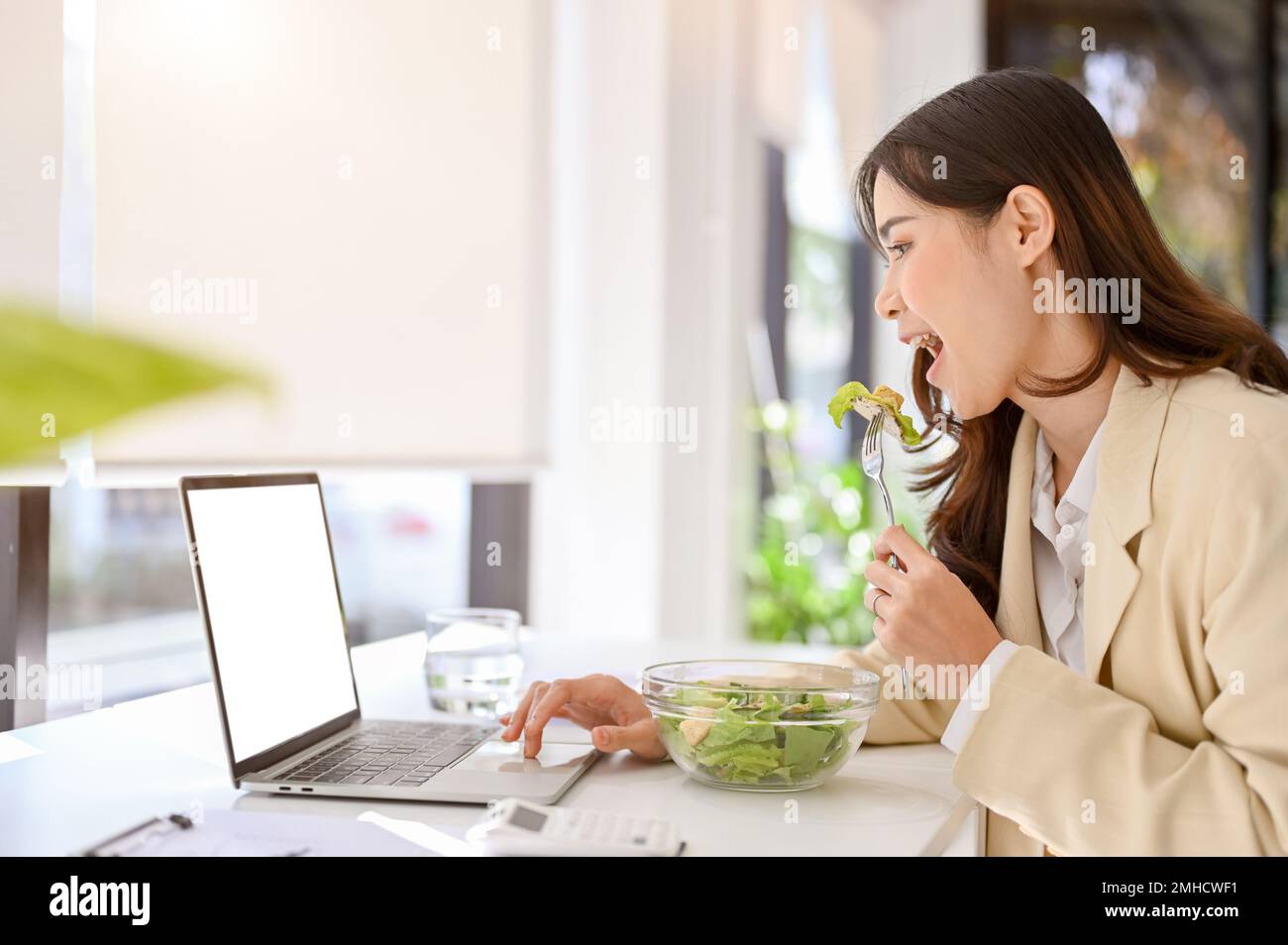 side view of a busy and hungry millennial asian businesswoman eating salad, having lunch at her desk while working on her project on laptop. busy urba Stock Photo