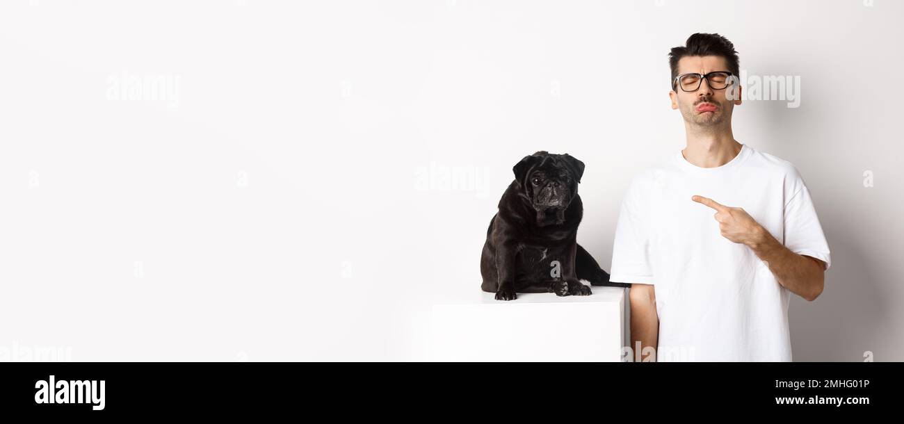 Sad and gloomy pet owner pointing at his black pug dog and sobbing, standing upster against white background Stock Photo