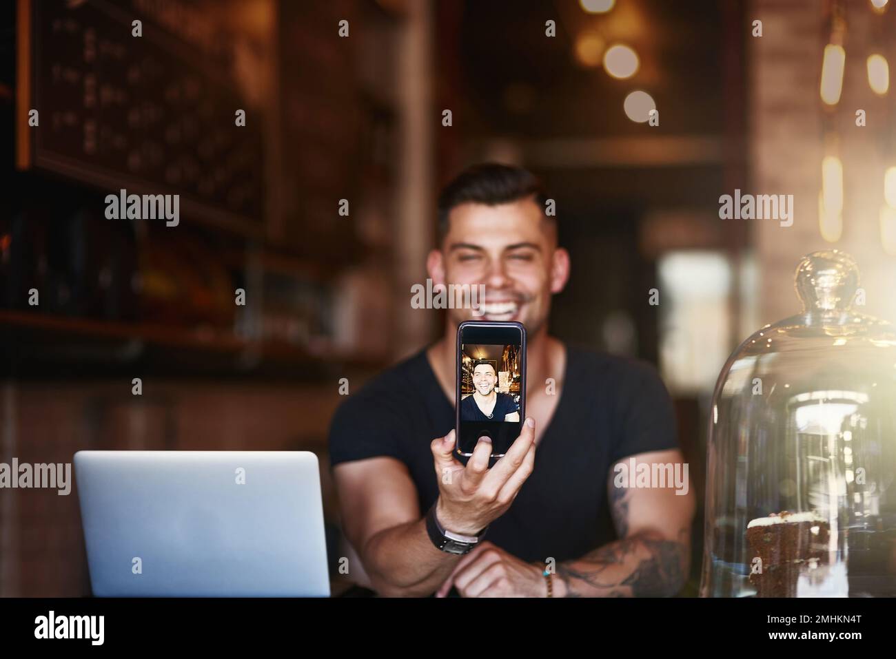 His smile sells a lot of coffee. a young man using a phone and laptop while working in a coffee shop. Stock Photo