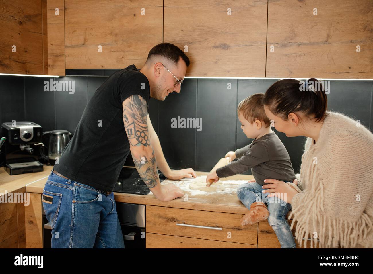 Happy parents with baby son together roll out raw dough with rolling pin on kitchen tabletop, side view. Family cooking. Stock Photo