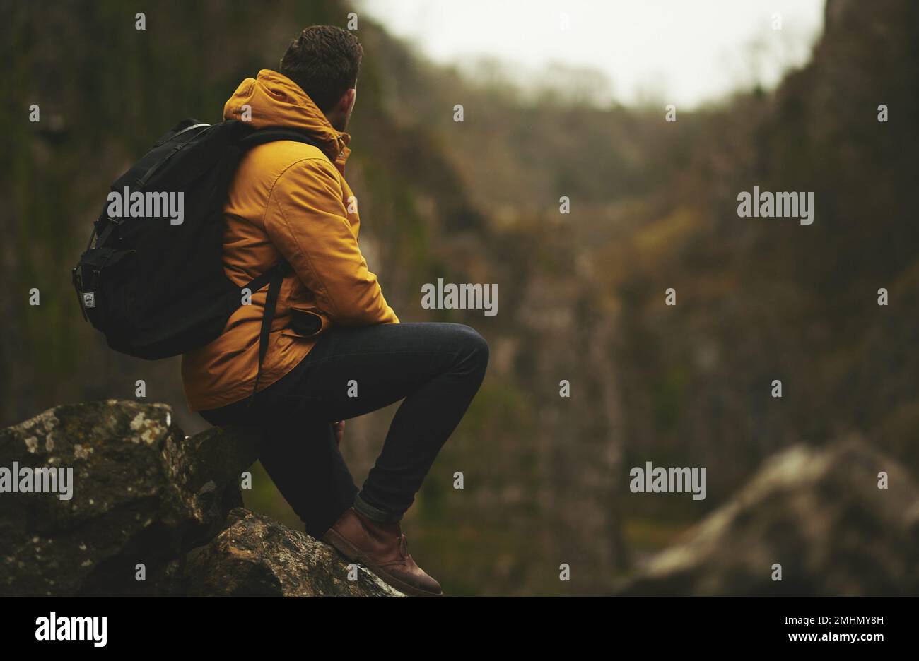 Hiker with backpack sitting on top of the rock mountain looking at the amazing environment. Hiker with a yellow jacket seated on a mountain edge. Stock Photo