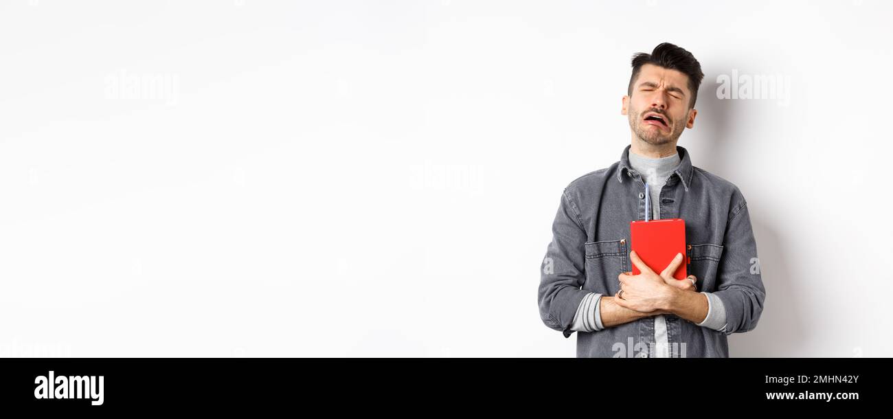 Sad cryig man holding red diary and sobbing, miserable guy carry journal with him, standing against white background Stock Photo