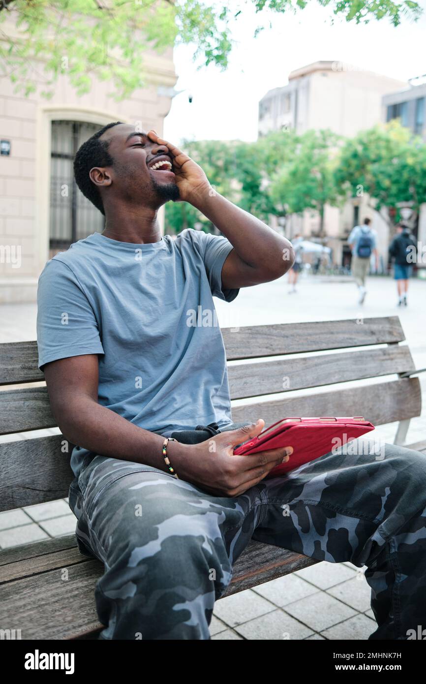 Man smiling while using a digital tablet sitting on a bench on the street. Stock Photo