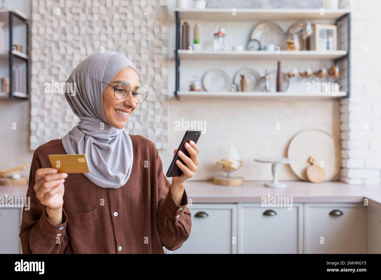Young smiling Arab woman in hijab using phone and credit card at home in kitchen. Do online shopping. Stock Photo