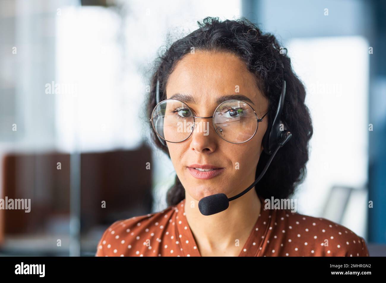 Close-up photo. Portrait of a young beautiful Latin American woman in a headset. Call center, service operator, hotline, support and assistance line. He looks seriously into the camera. Stock Photo
