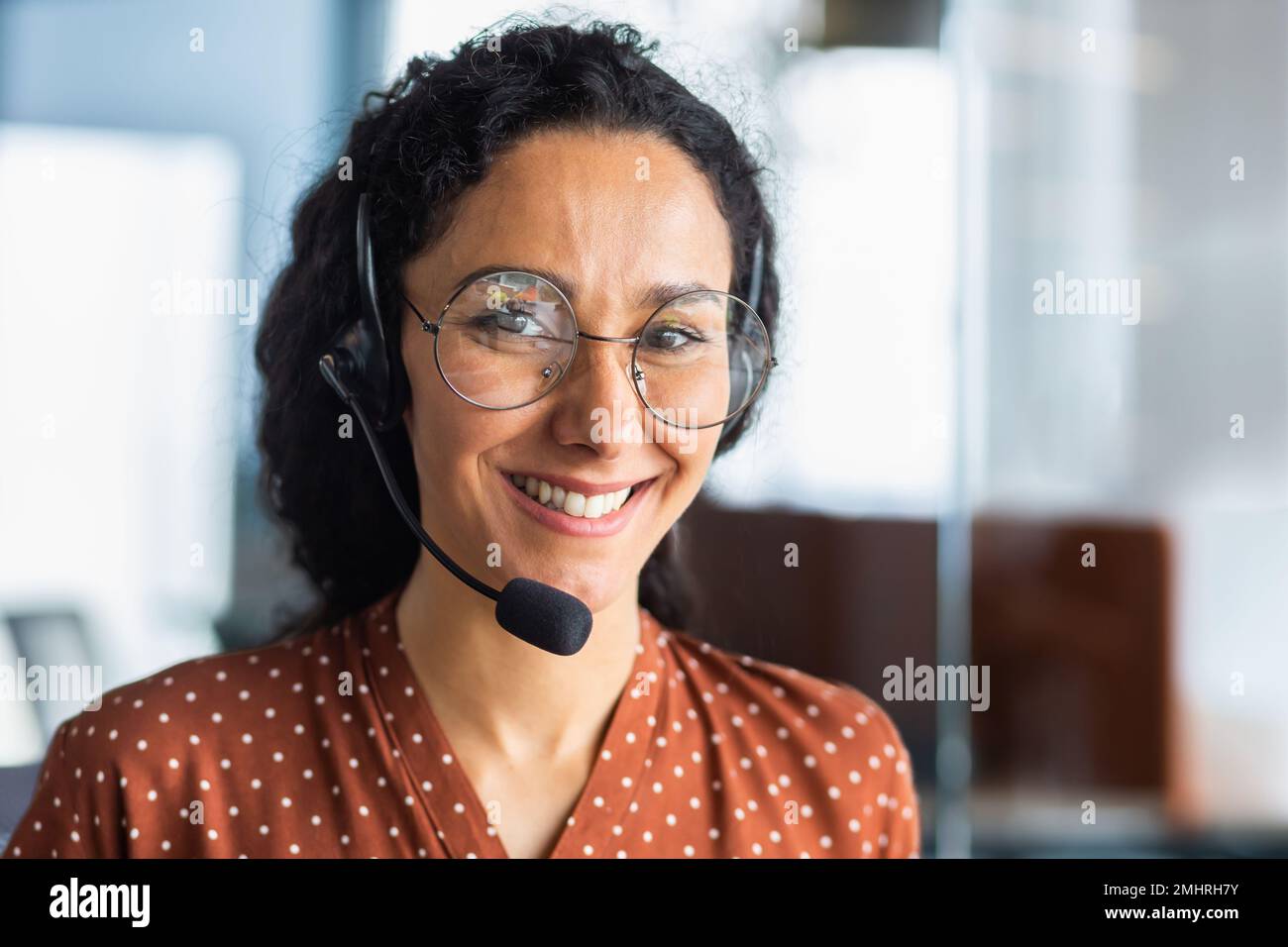 Close-up photo. Portrait of a young beautiful Latin American woman in a headset. Call center, service operator, hospital reception, support and assistance line. He looks at the camera, smiles. Stock Photo