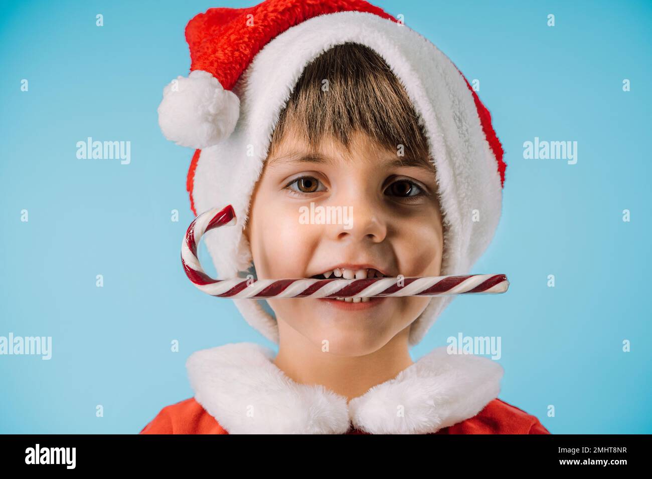 Funny boy in Santa Claus hat and sweet caramel cane on blue studio background. Christmas celebration. Happy childhood, kid, lovely son. High quality p Stock Photo