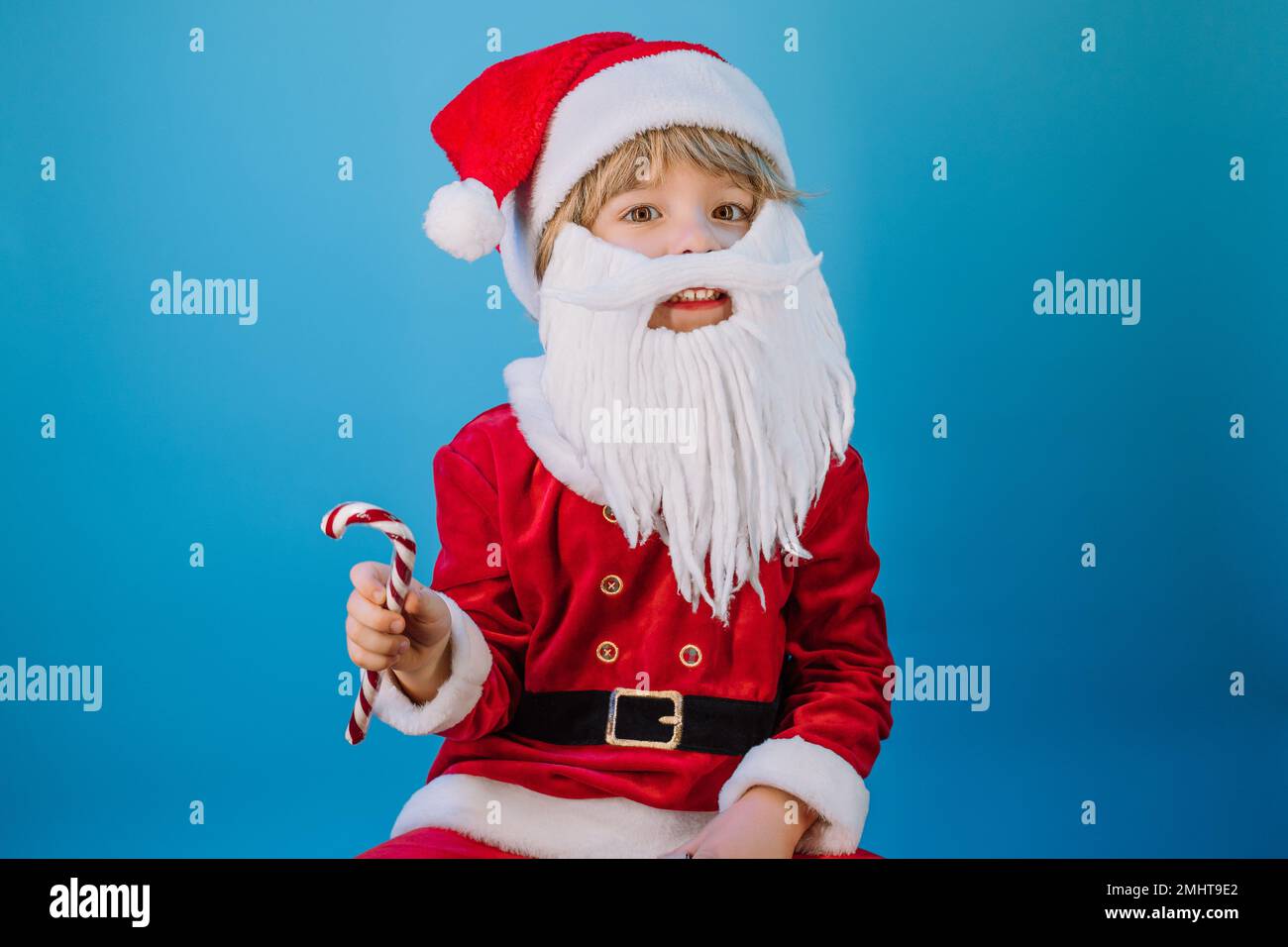 Funny boy in Santa Claus hat and sweet caramel cane on blue studio background. Christmas celebration. Happy childhood, kid, lovely son. High quality p Stock Photo