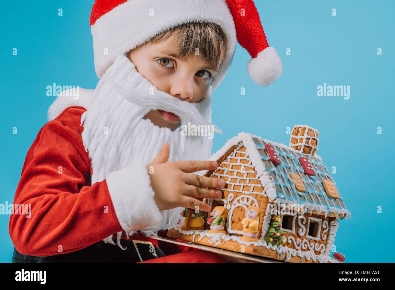 Cute little child in Santa Claus costume sitting with gingerbread house on blue background. Christmas celebration. Happy childhood, kid, lovely son. H Stock Photo