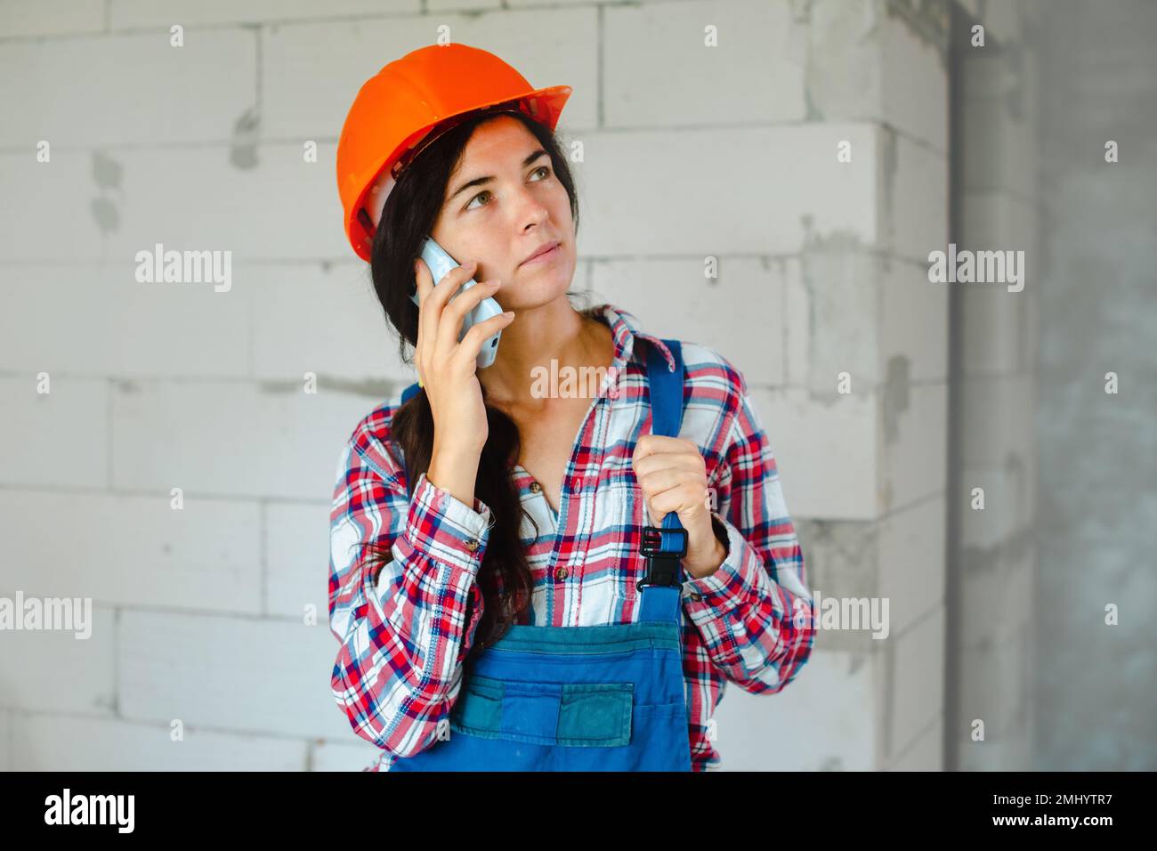 concentrated worker woman in blue overall having phone conversation during break from work at construction site indoors Stock Photo