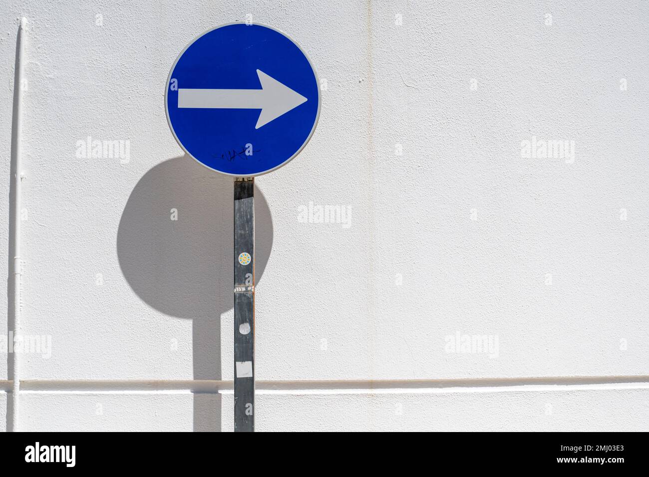 A blue one way traffic sign against a white wall with strong shadows in bright sunlight in a street in Portugal Stock Photo