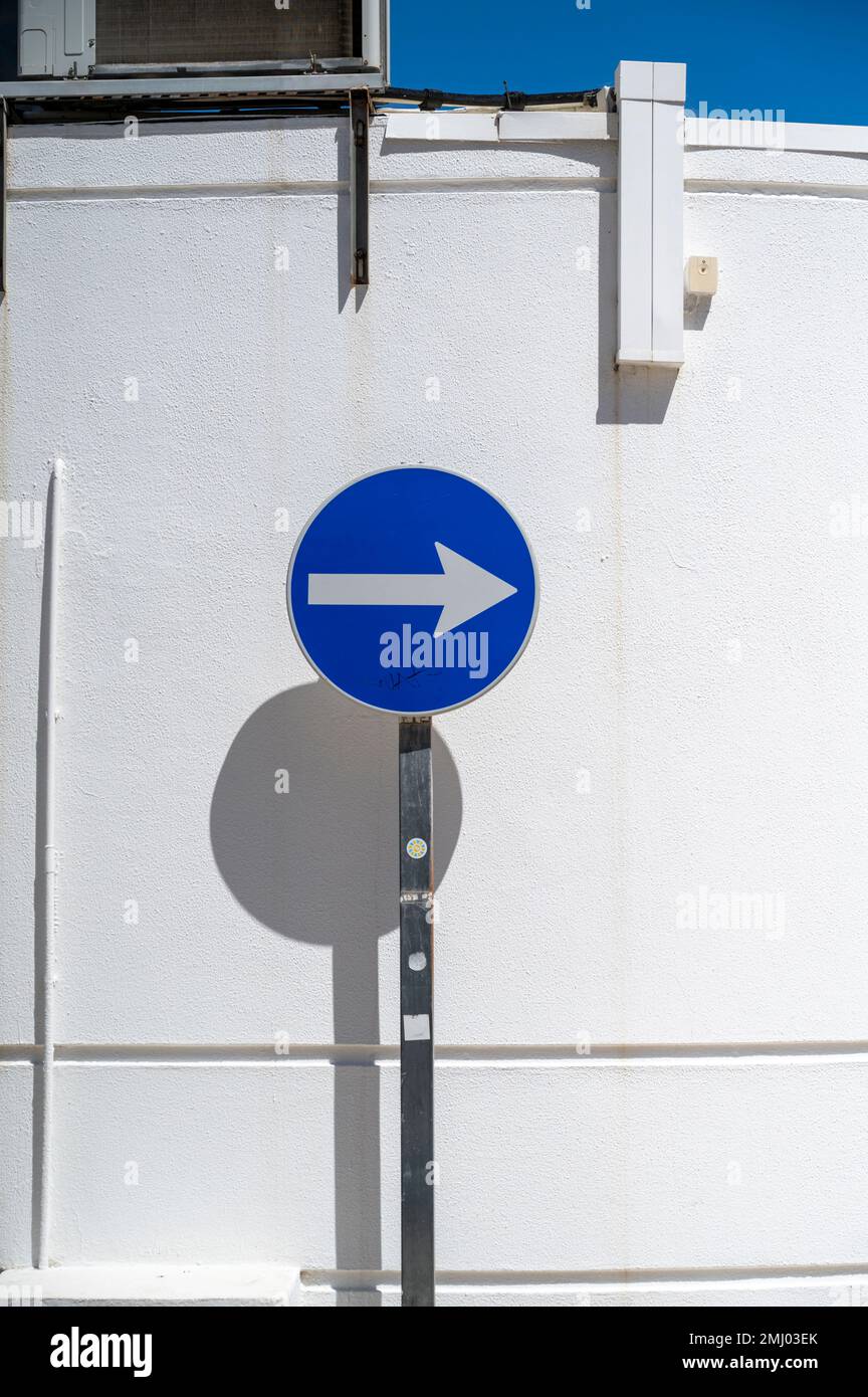 A blue one way traffic sign against a white wall with strong shadows in bright sunlight in a street in Portugal Stock Photo