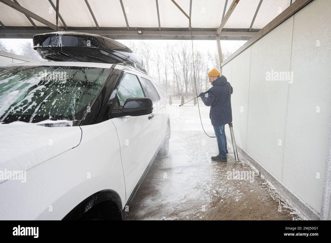 A man holds a foam sprayer from which foam flies out onto a car. A car at a  self service car wash. Stock Photo by romanchoknadii