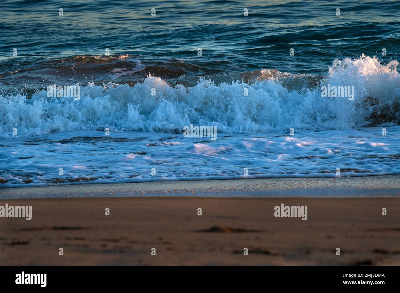 The ocean's waves crash on a sandy beach at sunset. Stock Photo