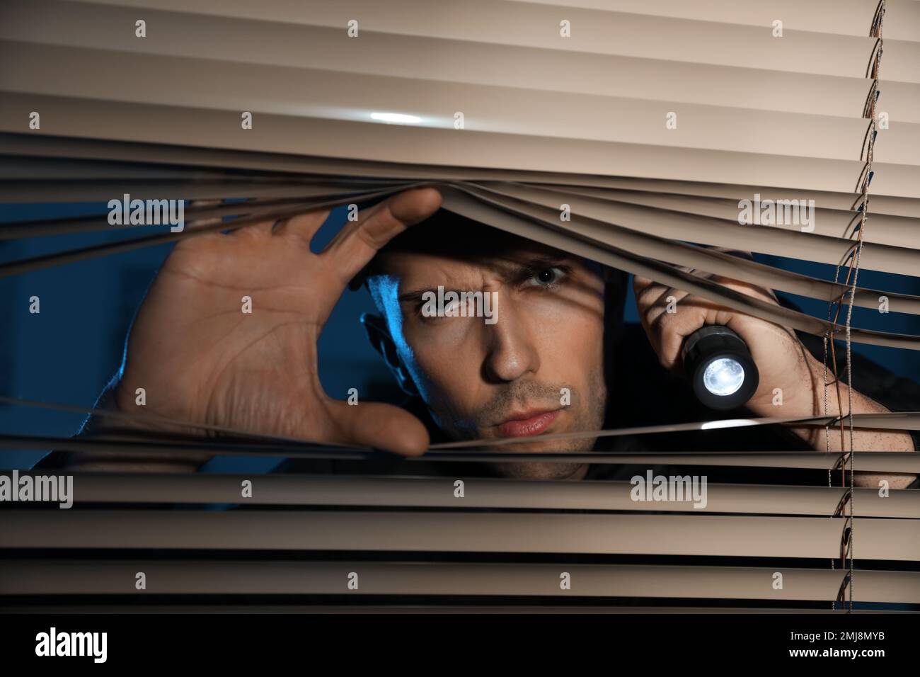 Male security guard with flashlight looking through window blinds in