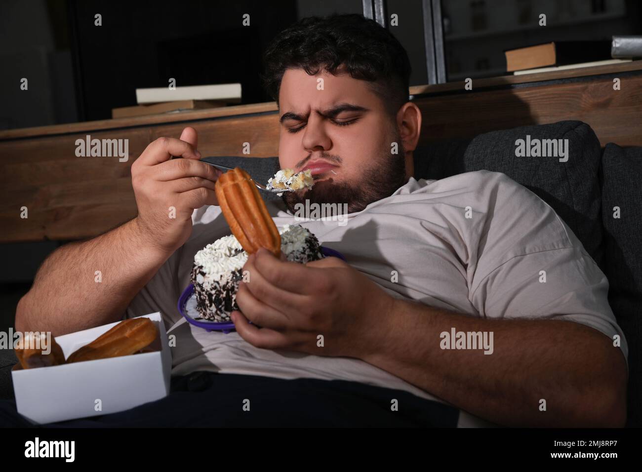Depressed overweight man eating sweets in living room at night Stock