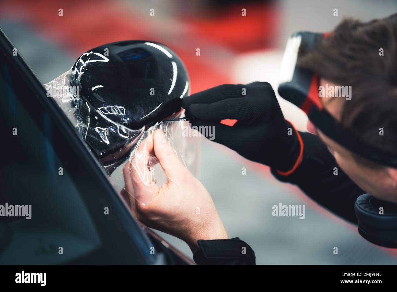 Close-up shot of man wearing glove on one hand applying PPF Paint Protection Film to side mirror of black car. Professional car detailing. Horizontal indoor shot. High quality photo Stock Photo