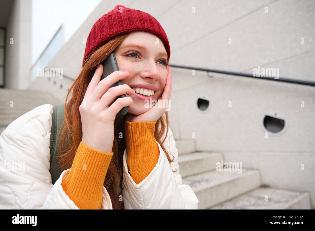 Beautiful smiling redhead female model, sits on street and talks on mobile phone, uses smartphone app to call abroad, laughing during telephone Stock Photo