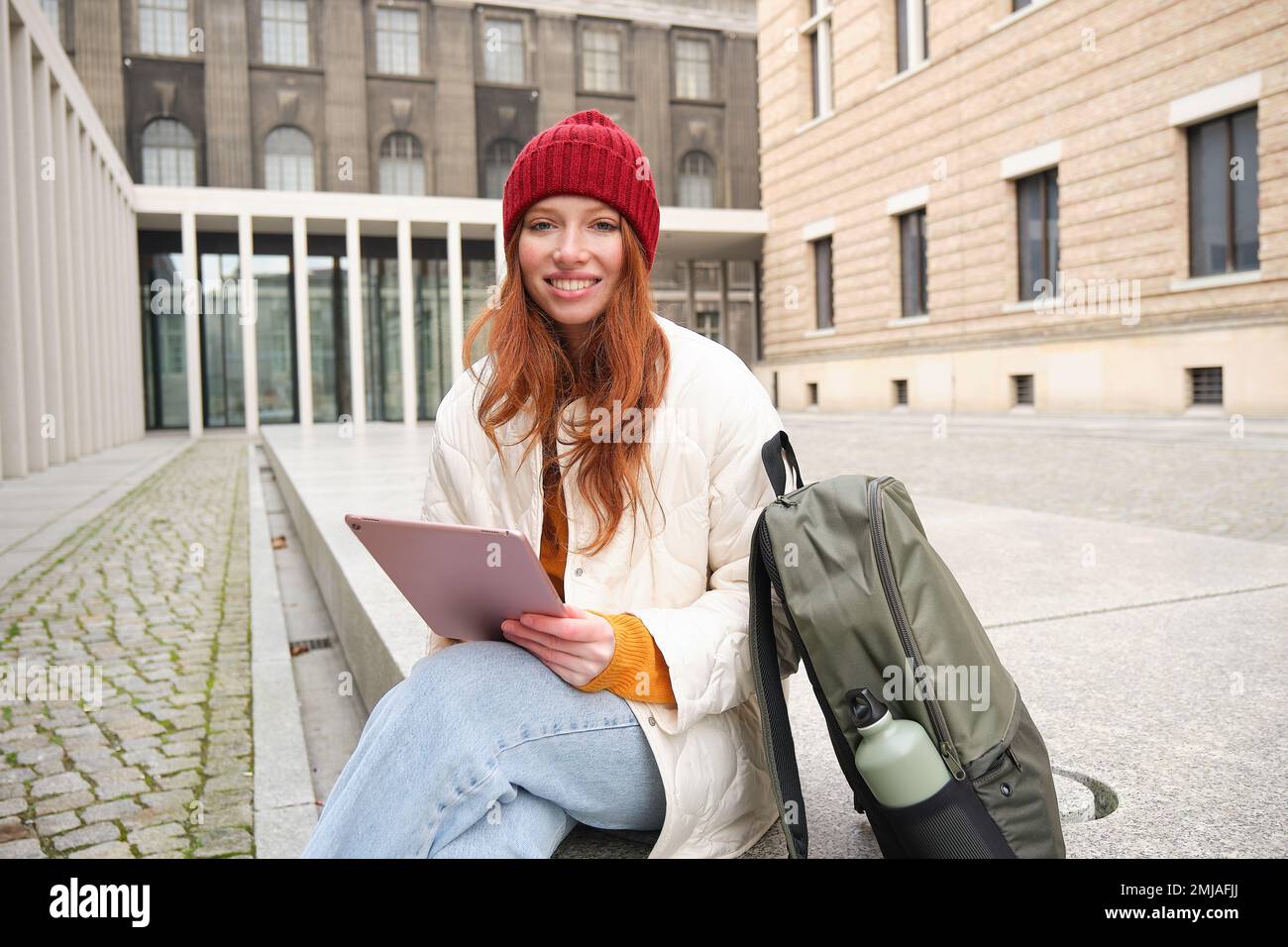 Beautiful redhead woman in red hat, sits with backpack and thermos, using digital tablet outdoors, connects to wifi, texts message, books tickets Stock Photo