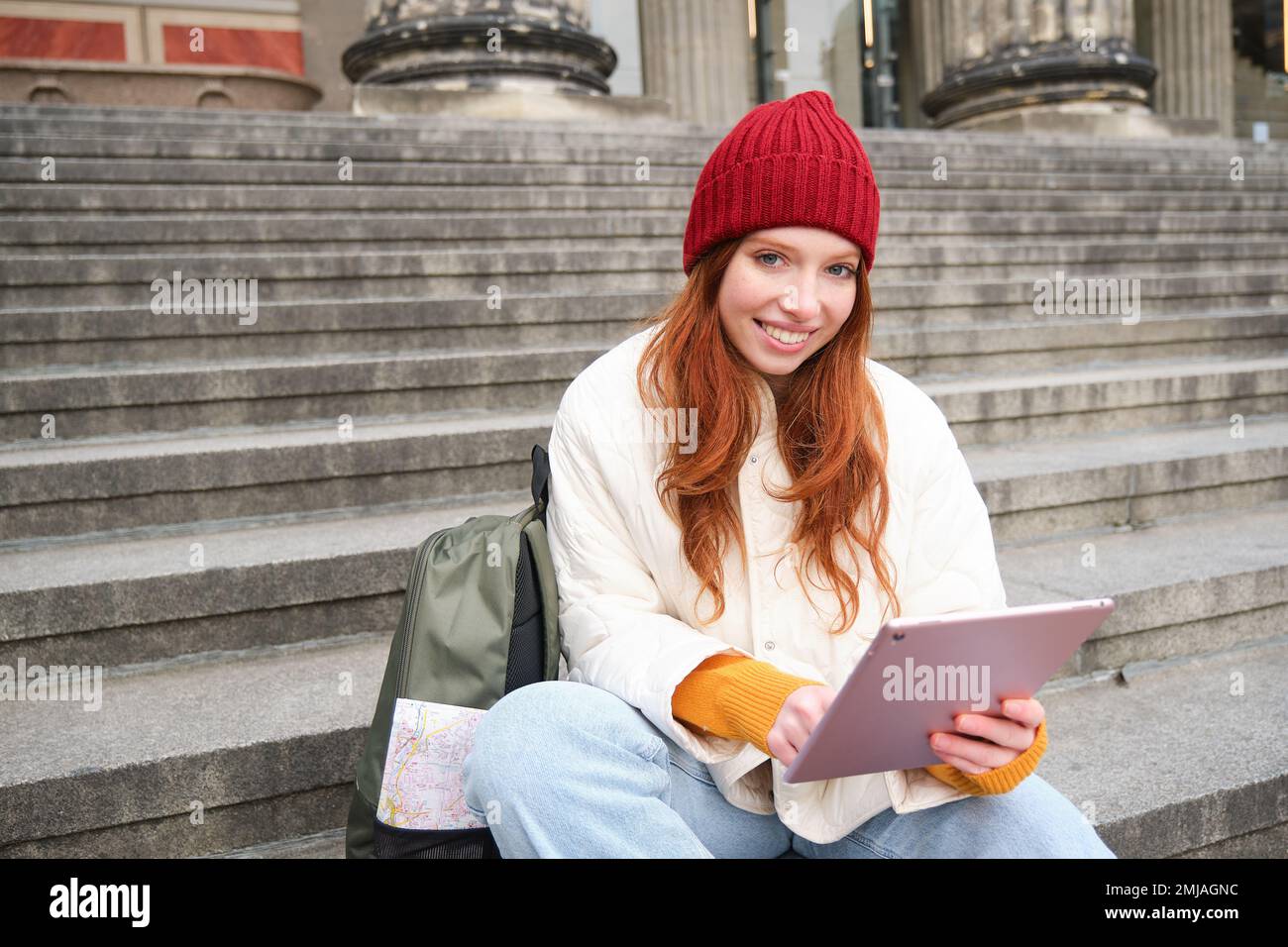 Beautiful young modern girl with red hair, holds digital tablet, sits on stairs near museum and connects public internet, sends message on gadget app Stock Photo