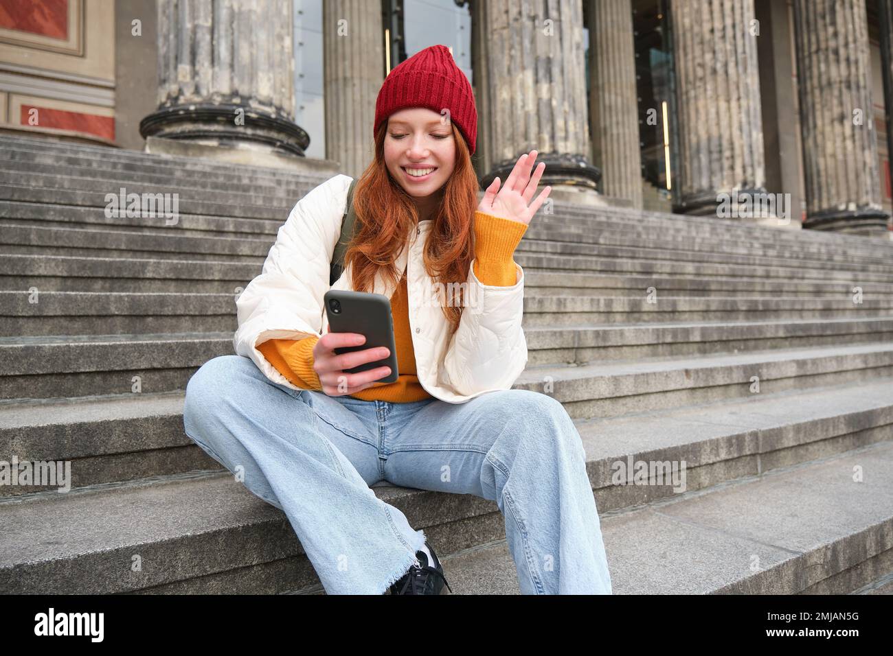 Redhead female tourist talks on mobile phone, video chatting with friends, says hi and waves hand at smartphone camera Stock Photo